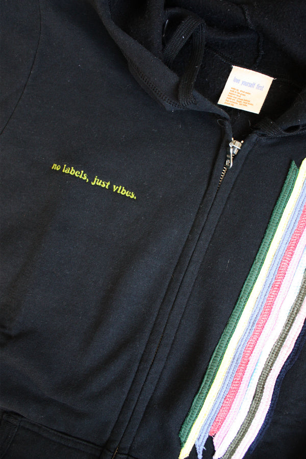 upcycled black zip up hoodie with colourful fabric stripes on the front, repurposed in canada, detail shot of 'no labels, just vibes.' embroidery